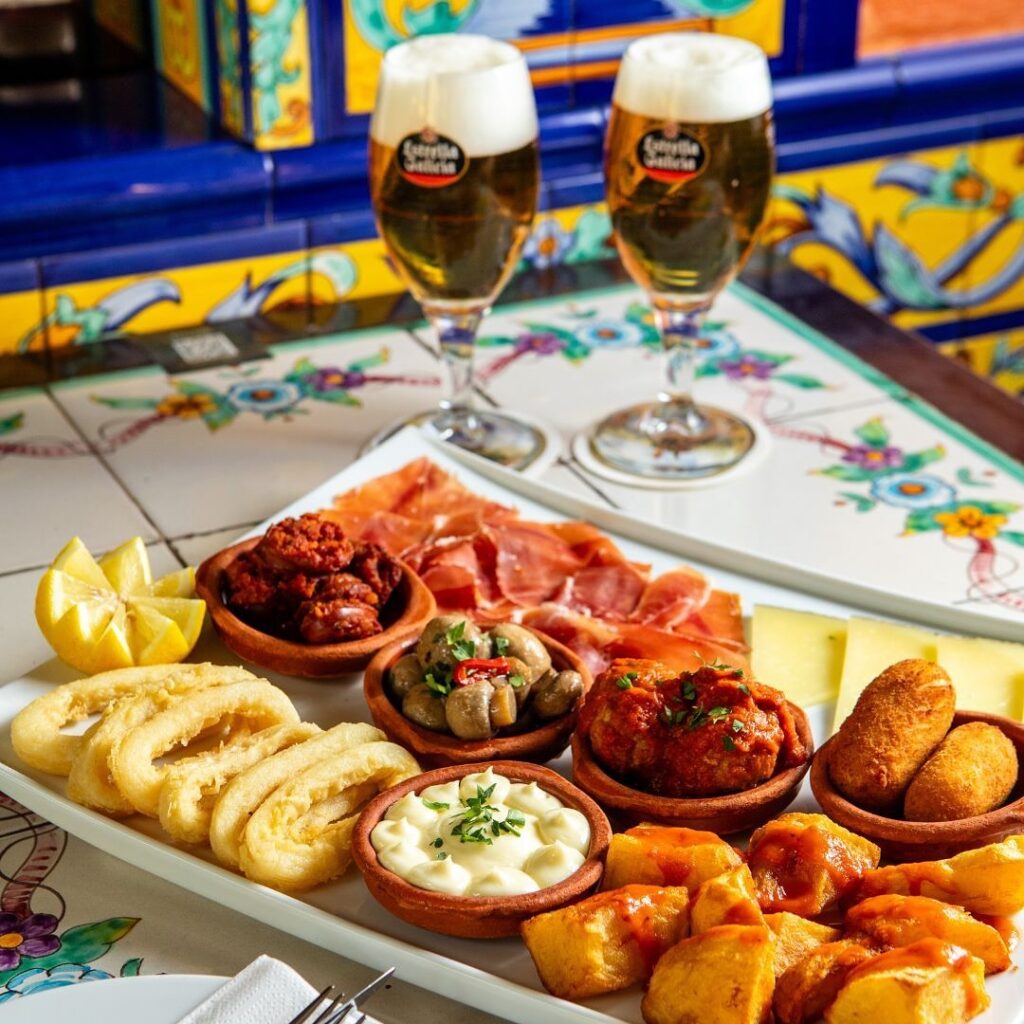 Ein spanischer Tapasteller mit zwei Gläsern Estrella Galicia Bier, serviert auf einem bunten Mosaiktisch im Vivolo Olé in Berlin.